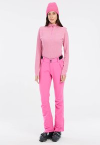 Roze patroon langarmtop met een ritskraag, gecombineerd met effen roze skibroeken. Model draagt zwarte skiboots. Achtergrond is wit.