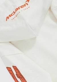 Bela kapuca s oranžnim logotipom "McLaren Formula 1 Team" in poudarkom. Gladka tekstura z modernim dizajnom in strukturirano kapuco.