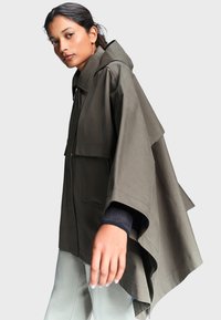 Femme portant une veste cape oversize vert olive à larges manches et un pantalon clair, regardant vers la caméra.