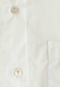 Dettaglio di una camicia bianca con bottoni, realizzata in un tessuto morbido, due bottoni in madreperla e una tasca applicata sulla parte frontale.