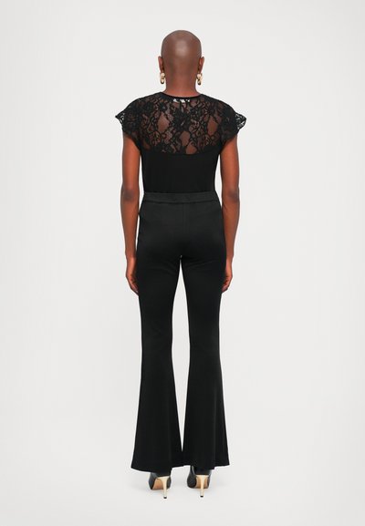 Femme à la tête rasée portant un haut en dentelle noire à manches courtes, un pantalon noir à taille haute et évasé, des boucles d'oreilles créoles dorées et des talons hauts noirs.