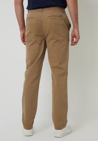 Pantalones de algodón beige de corte recto, con dos bolsillos traseros y una etiqueta de logo visible. La cintura cuenta con trabillas para cinturón y se estiliza con zapatillas blancas.