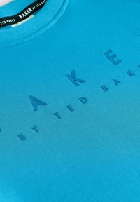 Niebieska bluza z teksturowym nadrukiem graficznym słowa "BAKER" napisanym dużymi literami oraz "by TED BAKER" w mniejszej czcionce.