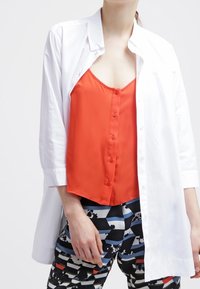 Chemise blanche boutonnée superposée sur un débardeur en satin rouge avec des boutons. Le bas présente un motif géométrique noir, bleu et rouge.