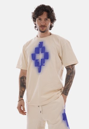 Uomo con capelli ricci e tatuaggi che indossa una t-shirt beige oversize e pantaloni con disegni geometrici blu a forma di croce, in piedi con una mano in tasca.