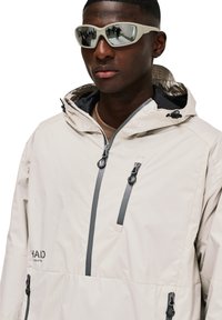 Helle beige Kapuzenjacke mit strukturiertem Finish, ausgestattet mit Reißverschlusstaschen auf der Brust und grauen Akzenten. Aufgesetzte Kapuze und verstellbare Kordeln.