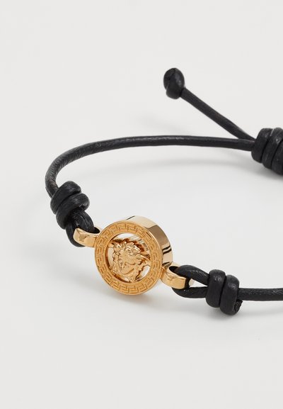 Bracciale in pelle nera con medaglione rotondo in oro caratterizzato da un motivo di leone e un motivo a chiave greca. Regolabile con estremità annodate.