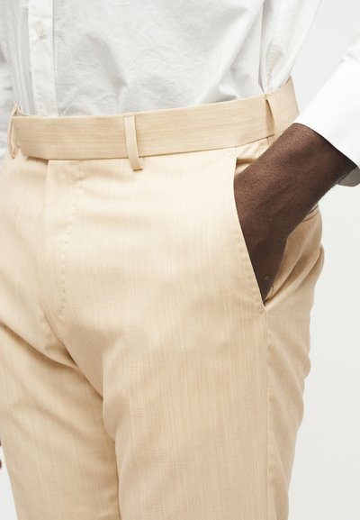 Άτομο με σκουρόχρωμο δέρμα που φοράει μπεζ παντελόνι και λευκό πουκάμισο, με το ένα χέρι άνετα στημένο στην τσέπη του παντελονιού.