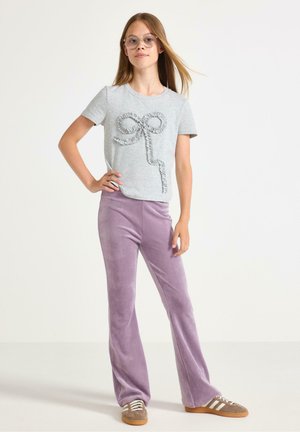Grijze T-shirt met een textuur strikdesign, gecombineerd met lavendelkleurige uitlopende broek van zacht materiaal, en bruine suède sneakers met witte strepen.