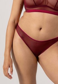 Kvinna i bourgogne transparenta underkläder, som visar torso, höfter och vänster arm med en blommatattoo på överarmen.