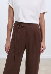 Bruna byxor med vida ben i texturerat tyg, framfållor och knytband i midjan. Matchade med en enkel vit t-shirt.