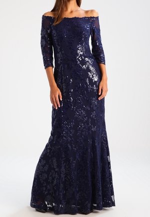 Femme portant une robe longue bleu marine, épaules dégagées, avec des manches en dentelle trois-quarts et des détails de paillettes florales.