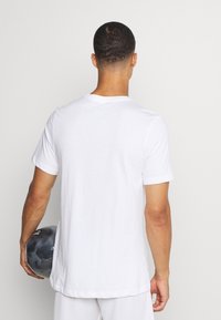 Uomo in maglietta bianca e pantaloni corta che tiene un pallone da calcio nero e grigio dietro la schiena, girato verso il basso su uno sfondo bianco uniforme.