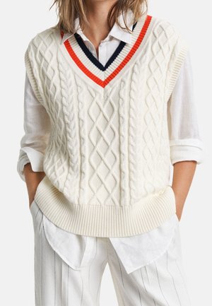 Femme portant un gilet sans manches en maille torsadée blanc avec un col en V rayé bleu marine et rouge, par-dessus une chemise blanche retroussée et un pantalon blanc à fines rayures.