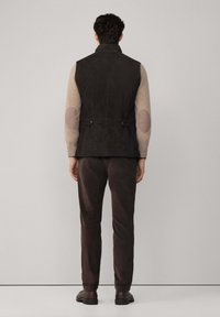 Gilet imbottito nero con colletto alto, toppe marroni sui gomiti e due bottoni in vita; abbinato a pantaloni e scarpe marroni scuri.