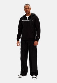 Felpa nera con zip e logo "Champion" bianco, abbinata a pantaloni cargo neri larghi e sneakers bianche con strisce nere.