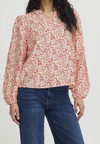 Blouse fleurie avec un col en V, présentant des motifs rouges et jaunes sur un fond rose clair. Manches longues bouffantes et coupe ample.