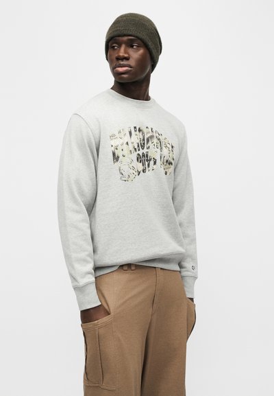 Άνδρας που φορά γκρι φούτερ με διακοσμητικό κείμενο, ανοιχτό καφέ παντελόνι με τσέπες cargo και χακί σκουφί, κοιτάζει προς τα πλάγια.