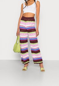 Pantalons en crochet à jambes larges avec des rayures multicolores en rose, vert, bleu et marron. Associés à un crop top blanc et un sac à main vert.