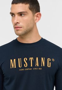 Tengerészkék, hosszú ujjú póló sárga felirattal: "MUSTANG" és szlogennel: "JEANS COMPANY SINCE 1932"; sima nyakkivágással és puha tapintással.
