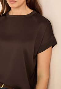 Hnědý top s krátkým rukávem, hladká struktura, volný střih s kulatým výstřihem, jemný manžet na rukávech, nošený s hnědým opaskem.