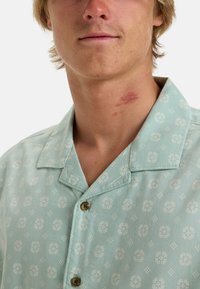 Camicia azzurro chiaro a maniche corte con un motivo floreale bianco. Tessuto morbido con colletto e due bottoni vicino al collo.