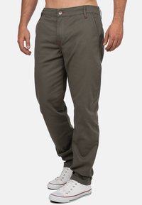 Pantalon vert olive foncé en mélange de coton avec une coupe droite. Caractéristiques : accents de couture rouge, braguette zippée et poches latérales. Porté avec des baskets blanches.