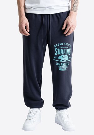 Muž v čiernych teplákových nohaviciach s tyrkysovým nápisom "Ocean Pacific Surfing Los Angeles" a bielymi teniskami stojaci s uvoľnenými rukami.