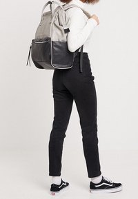 Personne portant un haut blanc à manches longues, un jean noir taille haute, des baskets noires et blanches, et portant un sac à dos gris et noir.