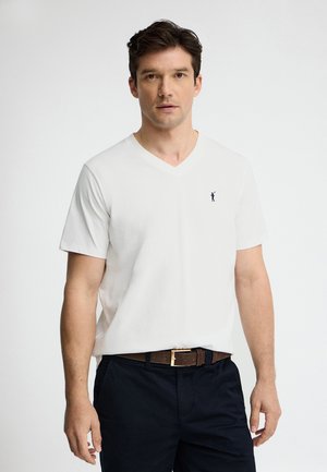 Mężczyzna w białej koszulce typu V-neck z małym czarnym logo oraz czarnych spodniach z brązowym paskiem, stojący na jednolitym białym tle.