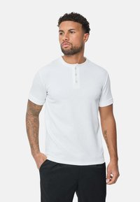 Chemise henley blanche à manches courtes en tissu texturé, avec une patte de boutonnage à trois boutons et un col rond. Portée avec un pantalon noir.