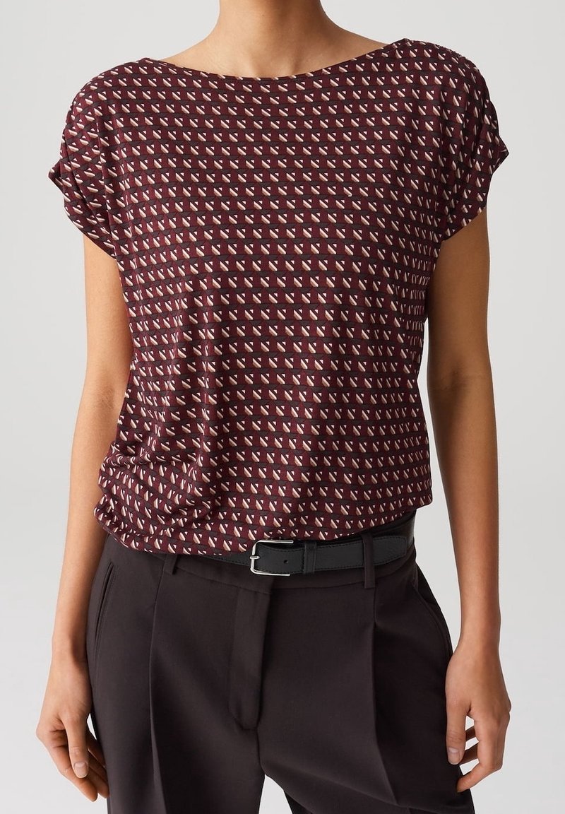 Bordeaux kortærmet bluse med et geometrisk mønster af små hvide trekanter. Blødt stof, afslappet pasform, med rund halsudskæring og oprullede ærmer.