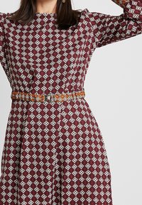 Une robe bordeaux présentant un motif floral blanc, avec des manches longues. Elle dispose d'une ceinture en cuir brun à motif avec une boucle en argent à la taille.
