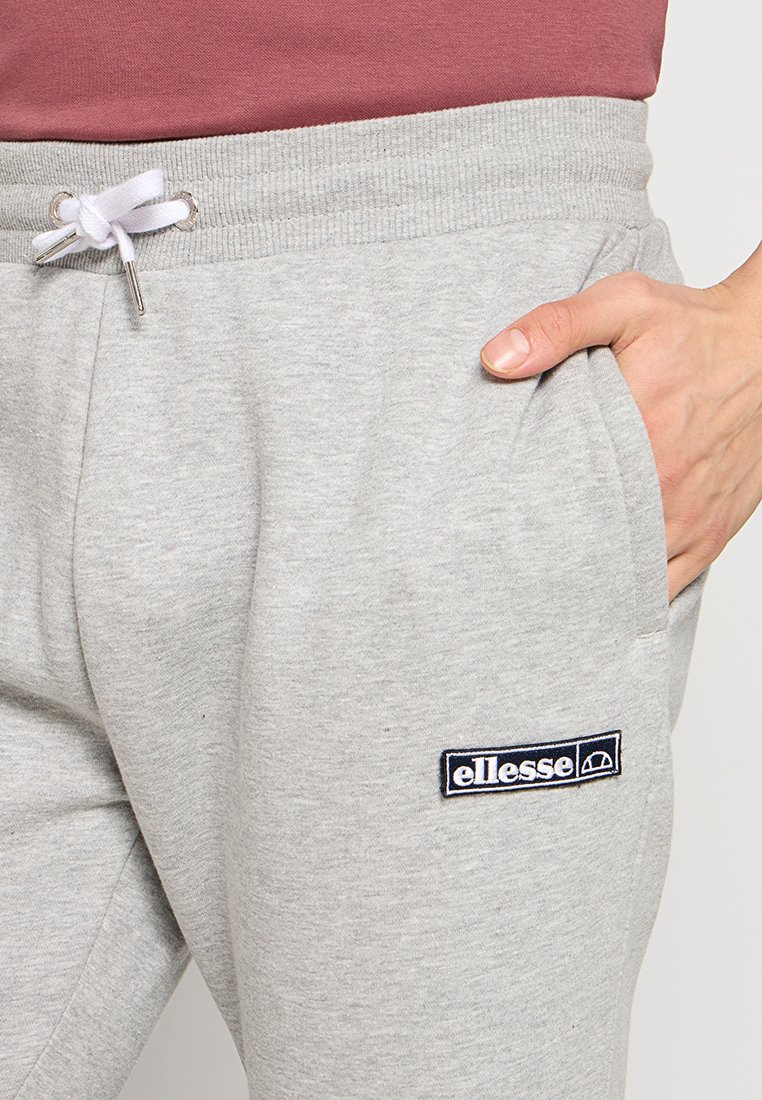 Pantalones de chándal grises hechos de tela suave, con una cintura elástica con cordón, dos bolsillos laterales y un parche de logo negro.