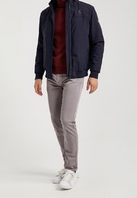 Navy blue zip-up jacket with high collar, paired with a burgundy sweater and light gray slim-fit pants. White sneakers complete the look.