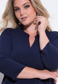 Dunkelblaues Oberteil mit V-Ausschnitt und 3/4-Ärmeln, das eine glatte Textur und ein dezentes Knopfdetail vorne aufweist. Die Ohrringe ergänzen den Look.