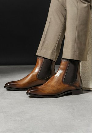 Brown leather Chelsea boots with elastic side panels, pointed toe, smooth texture, and contrast stitching, paired with light-colored trousers.