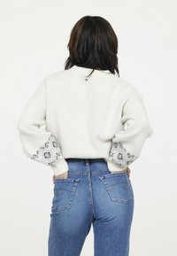 Pull en maille blanc avec des manches à motifs, coupe ample et poignets côtelés. Associé à un jean en denim bleu taille haute avec deux poches arrière.