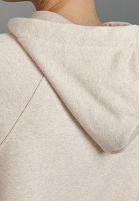 Sudadera color beige claro con un tejido suave y texturizado. Presenta una capucha redondeada con costuras visibles y un ligero solapamiento en el escote.