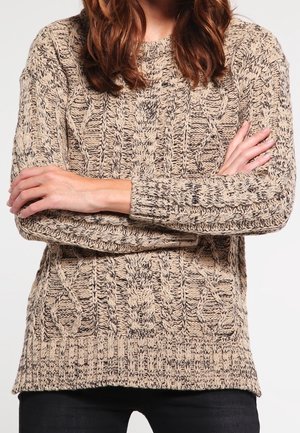 Personne aux longs cheveux bruns portant un pull beige à torsades, bras croisés, visible des épaules jusqu'aux hanches.