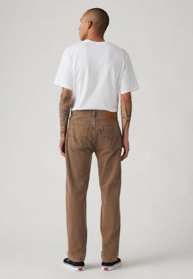 Man with tattoos wearing a white t-shirt, brown jeans, and black sneakers stands facing away against a plain light background.