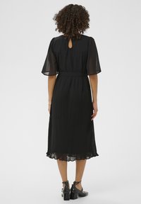 Rochie midi plisată neagră cu mâneci din plasă în formă de fluture și detalii cu tăietură în spate, combinată cu pantofi negri cu tocuri bloc.