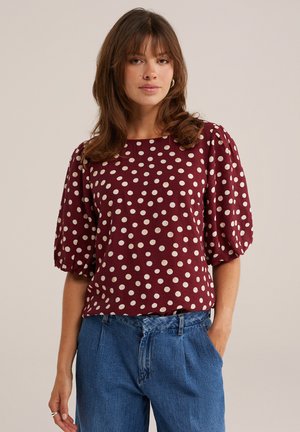 Mujer con cabello castaño que lleva una blusa burdeos con lunares blancos y mangas abullonadas y vaqueros azules de cintura alta con una mano en el bolsillo.