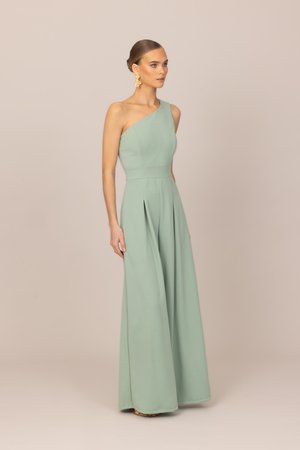 Frau trägt einen ärmellosen, einseitig schulterfreien, hellgrünen Jumpsuit mit weitem Bein und goldenen Statement-Ohrringen vor einem beigen Hintergrund.