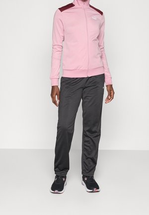 Person trägt eine hellrosa Reißverschlussjacke mit burgunderroten Akzenten auf den Schultern, schwarze Sporthose und schwarze Sneakers mit pinken Details.