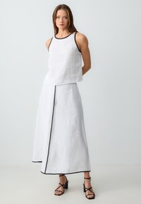 Top en lin blanc sans manches avec bordures noires associé à une jupe longue assortie avec une fente sur le devant et une couture d'accent noir. Sandales noires à lanières.
