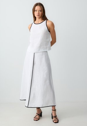 Weißes Leinen-Trägertop mit schwarzem Saum, kombiniert mit einem passenden langen Rock mit einem vorderen Schlitz und einer schwarzen Akzentnaht. Schwarze Riemchensandalen.