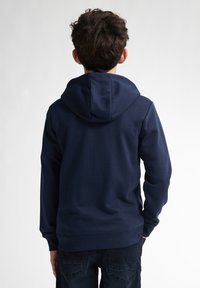 Hanorac cu glugă de culoare bleumarin, cu o croială lejeră, cu manșete ribbate și o textură netedă, asortat cu blugi închiși la culoare.