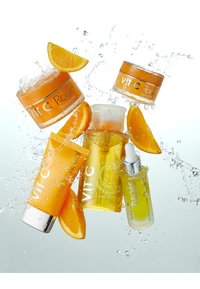 Fünf Hautpflegeprodukte in orangefarbener Verpackung, darunter ein Cremetiegel, ein Augen-Soufflé, eine SPF-Tube und zwei Pipettenflaschen, umgeben von Wasserspritzern und Orangenscheiben.