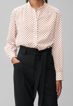 Kvinde iført en hvid skjorte med små røde blomstertryk, stoppet ned i højtaljede sorte bukser med et stofbælte.